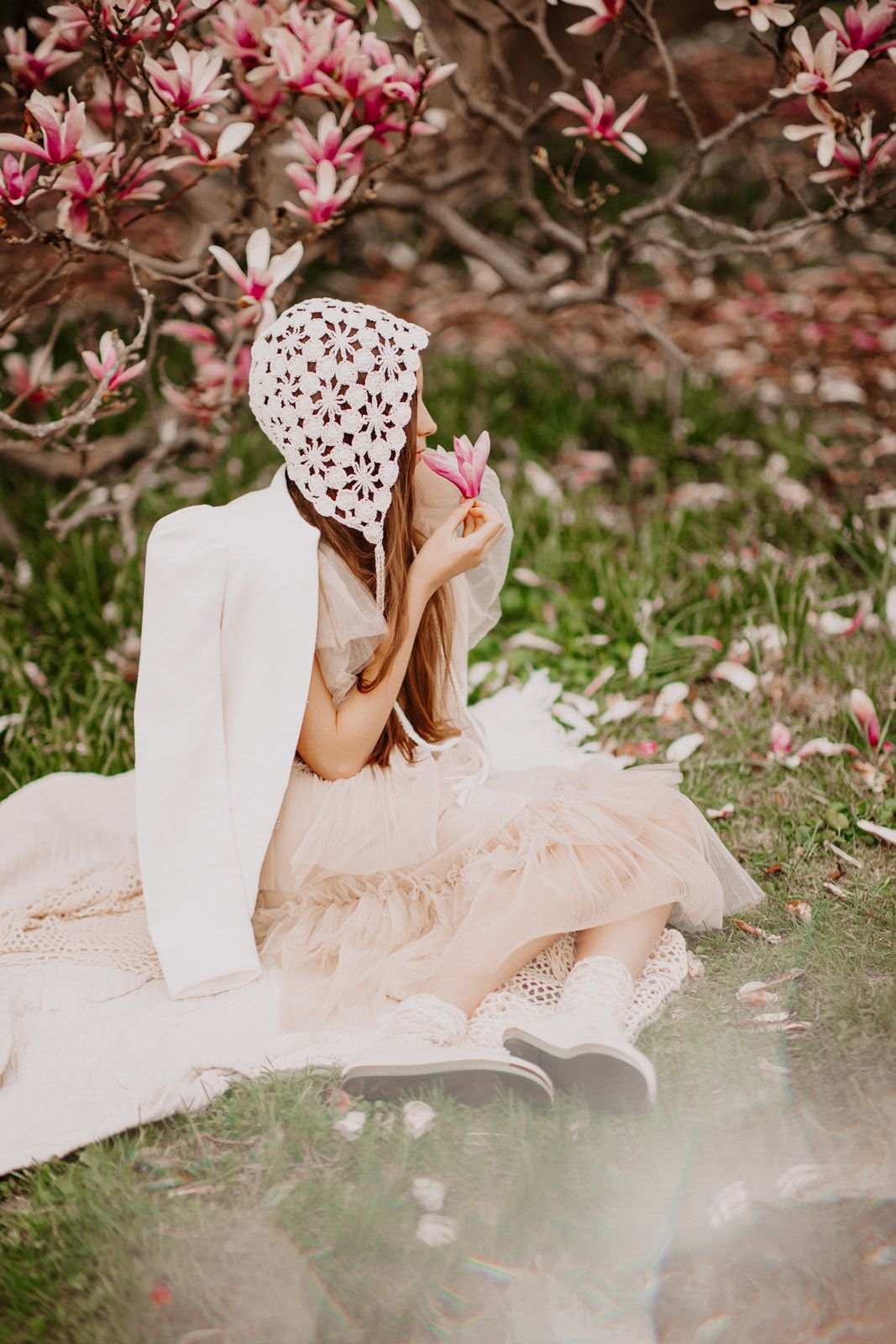 Young girl in white jacket and tulle skirt reads book under pink magnolia blossoms in spring garden.