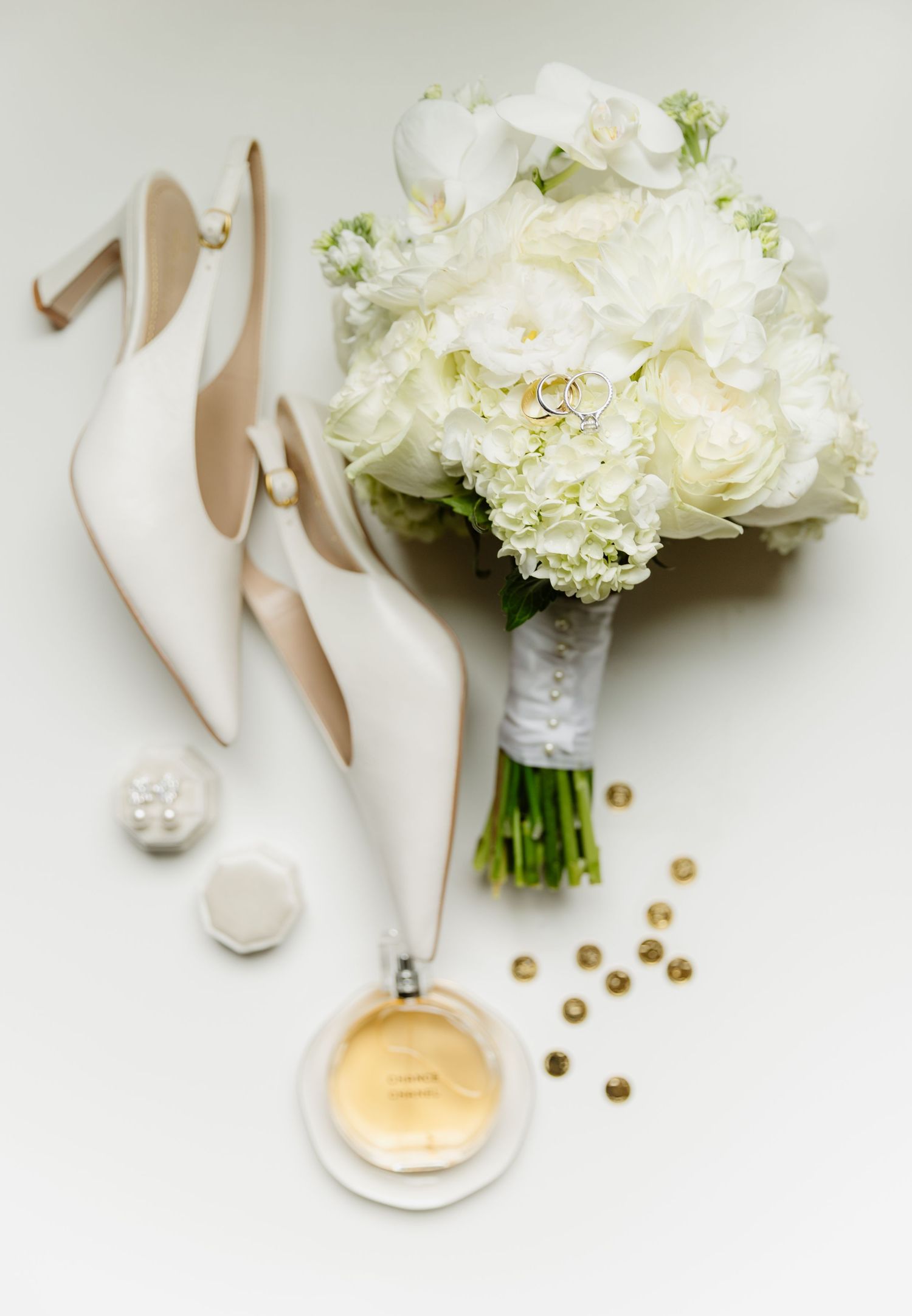 Elegant wedding flat lay featuring white bridal heels, a lush white floral bouquet, perfume, and gold accents.