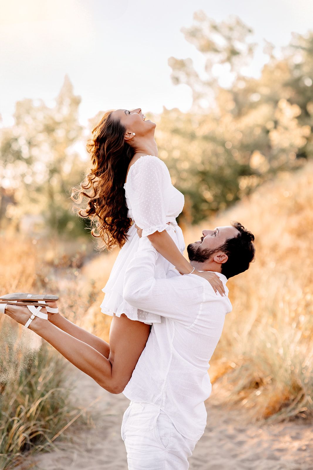 Couple in white outfits sharing a joyful moment outdoors, man lifting woman against golden sunlit nature backdrop.