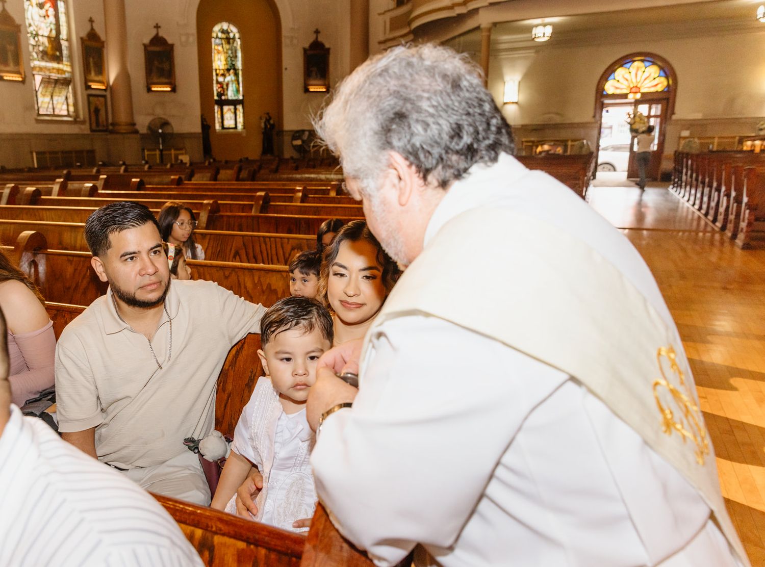 Catholic priest interacts with family and baby during a baptism ceremony inside a traditional church.