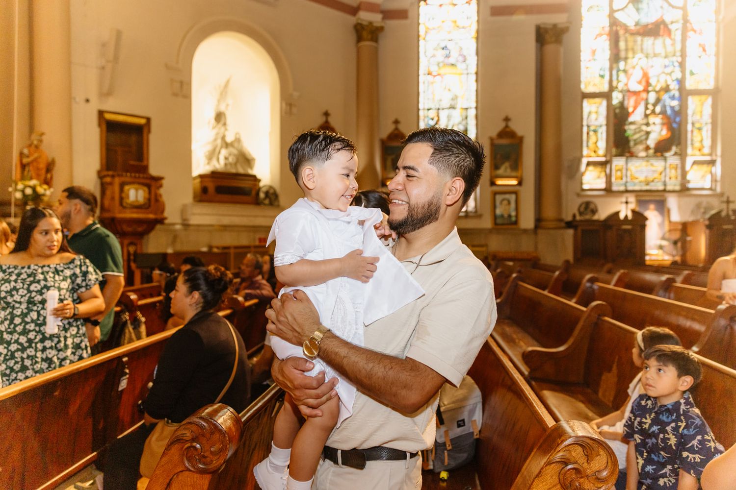 A father joyfully holds his baby during a church baptism ceremony, smiling warmly inside a beautiful cathedral.