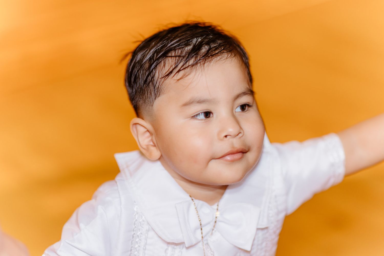 Cute toddler boy in white baptism outfit with bow tie against a bright orange background.