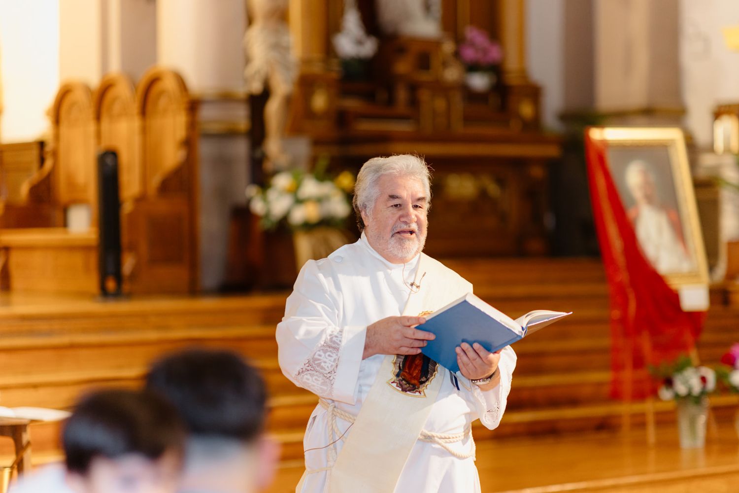 Catholic priest in white vestments reads from a blue book during a church ceremony at the altar.