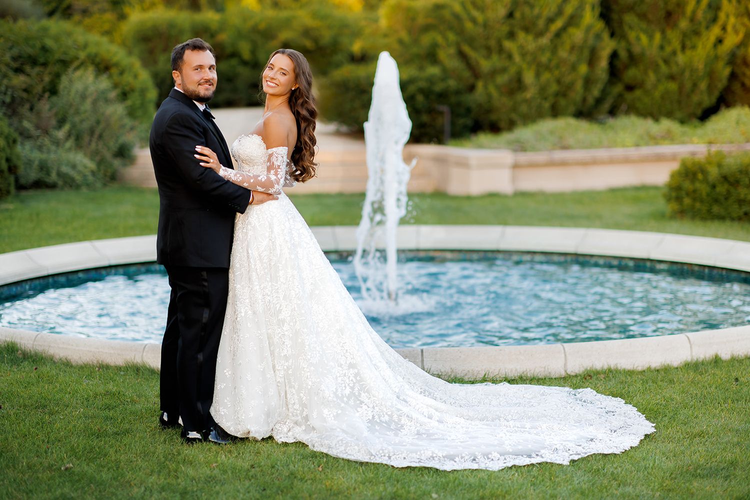 Newlywed couple poses by circular garden fountain, bride in lace off-shoulder gown with long train.