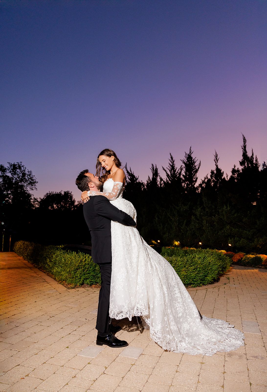 Groom lifts smiling bride in an elaborate lace wedding gown against a vibrant purple and blue dusk sky.