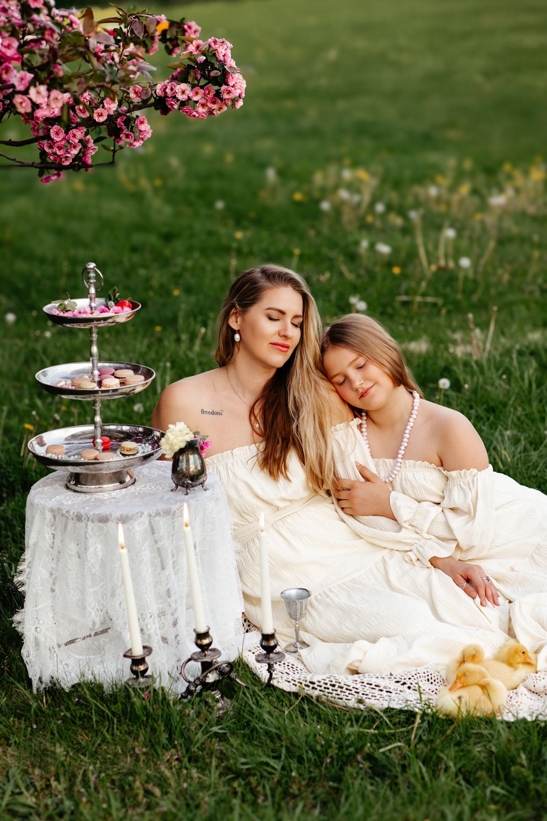 Mother and daughter in white dresses enjoy a picnic under blooming pink flowers on lush green grass.