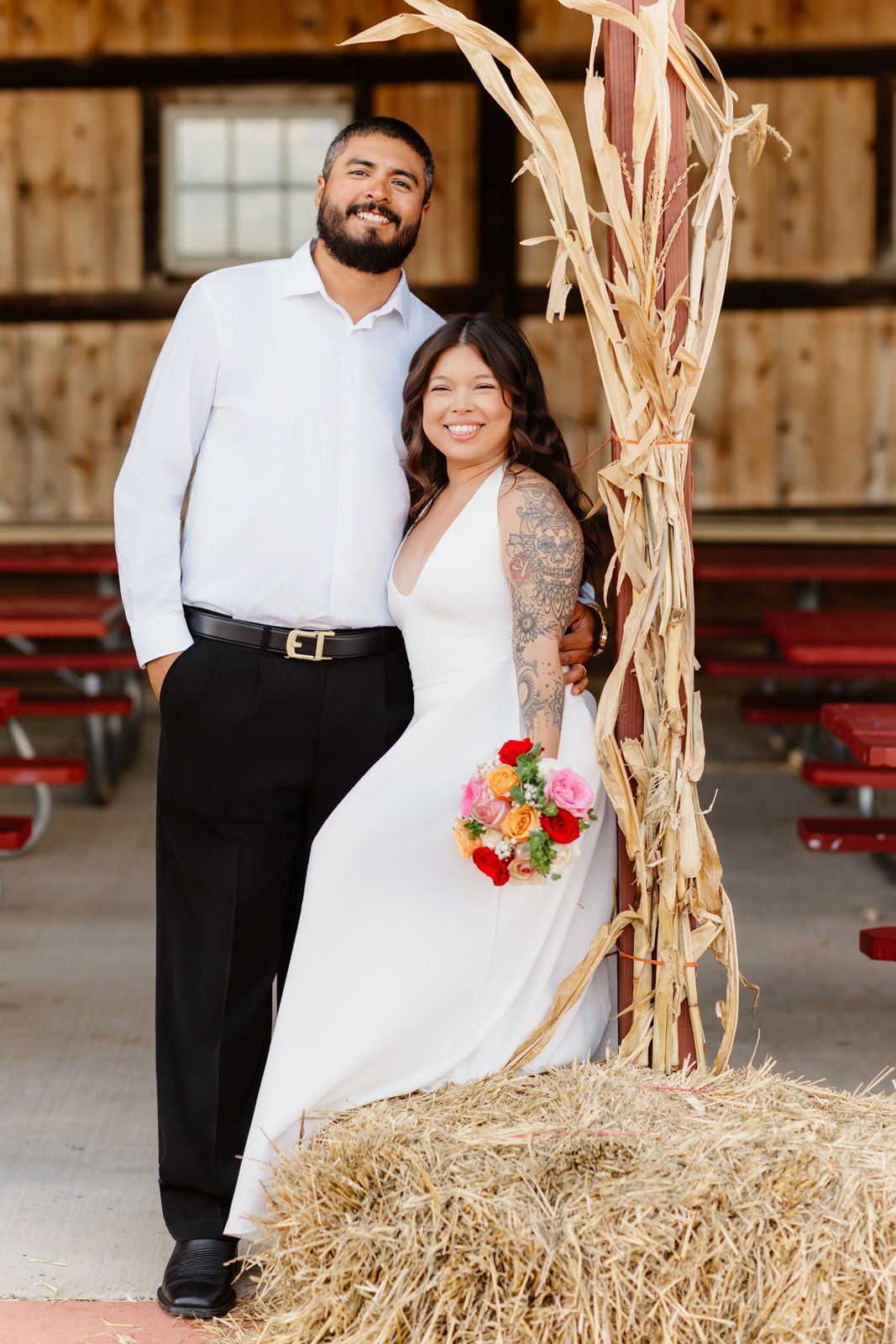 Smiling couple in wedding attire pose beside dried corn stalks on hay bale in rustic barn venue.