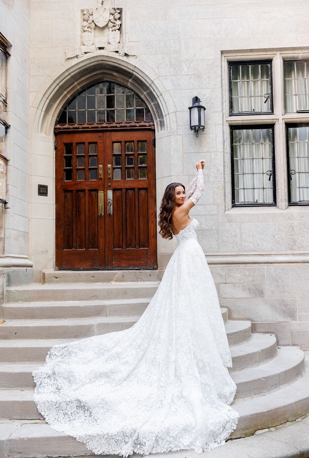 Bride in a long-sleeve lace wedding gown with a cathedral train poses on stone steps before ornate wooden doors.