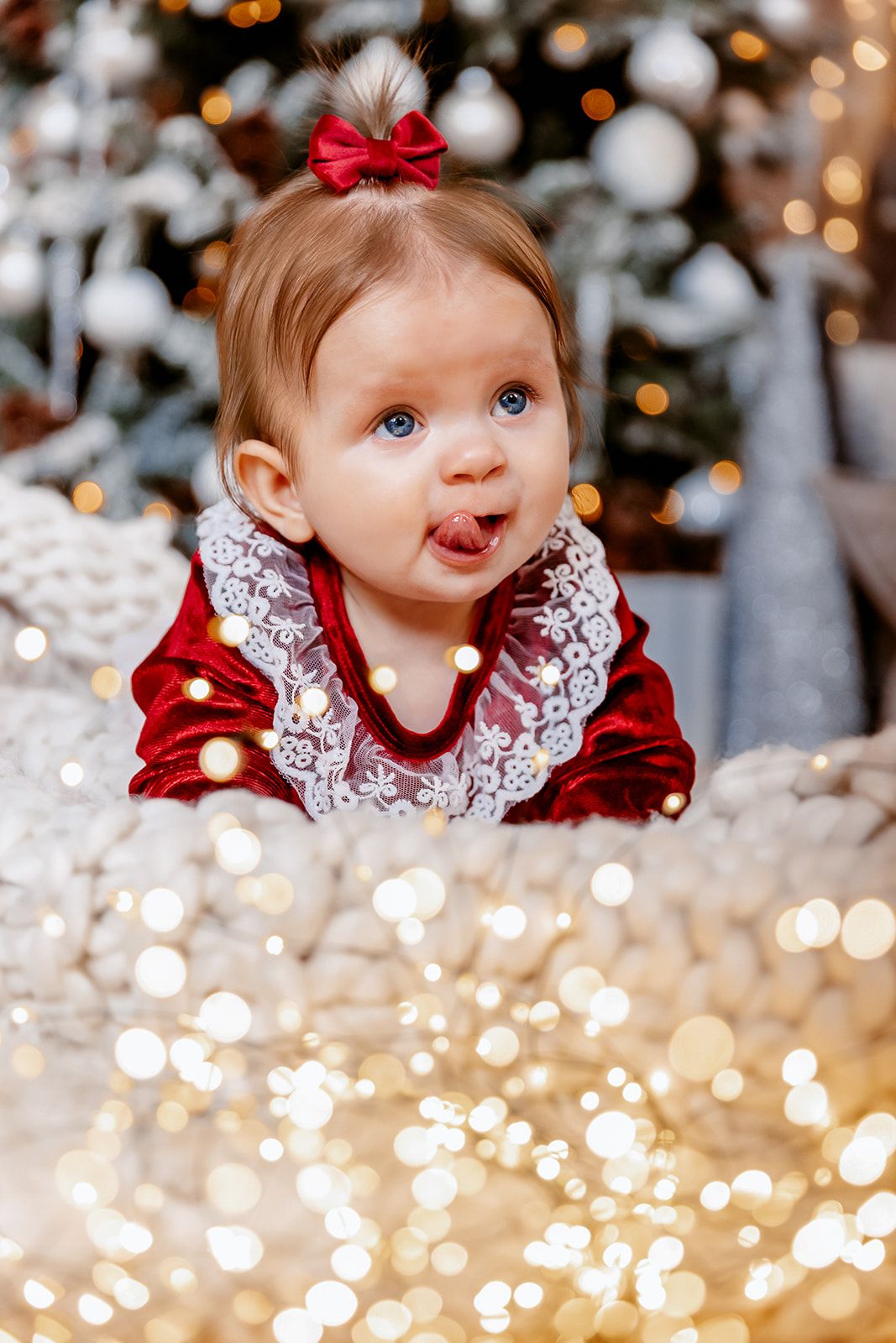 Adorable baby girl in red velvet dress sticking tongue out near decorated Christmas tree with golden bokeh lights.
