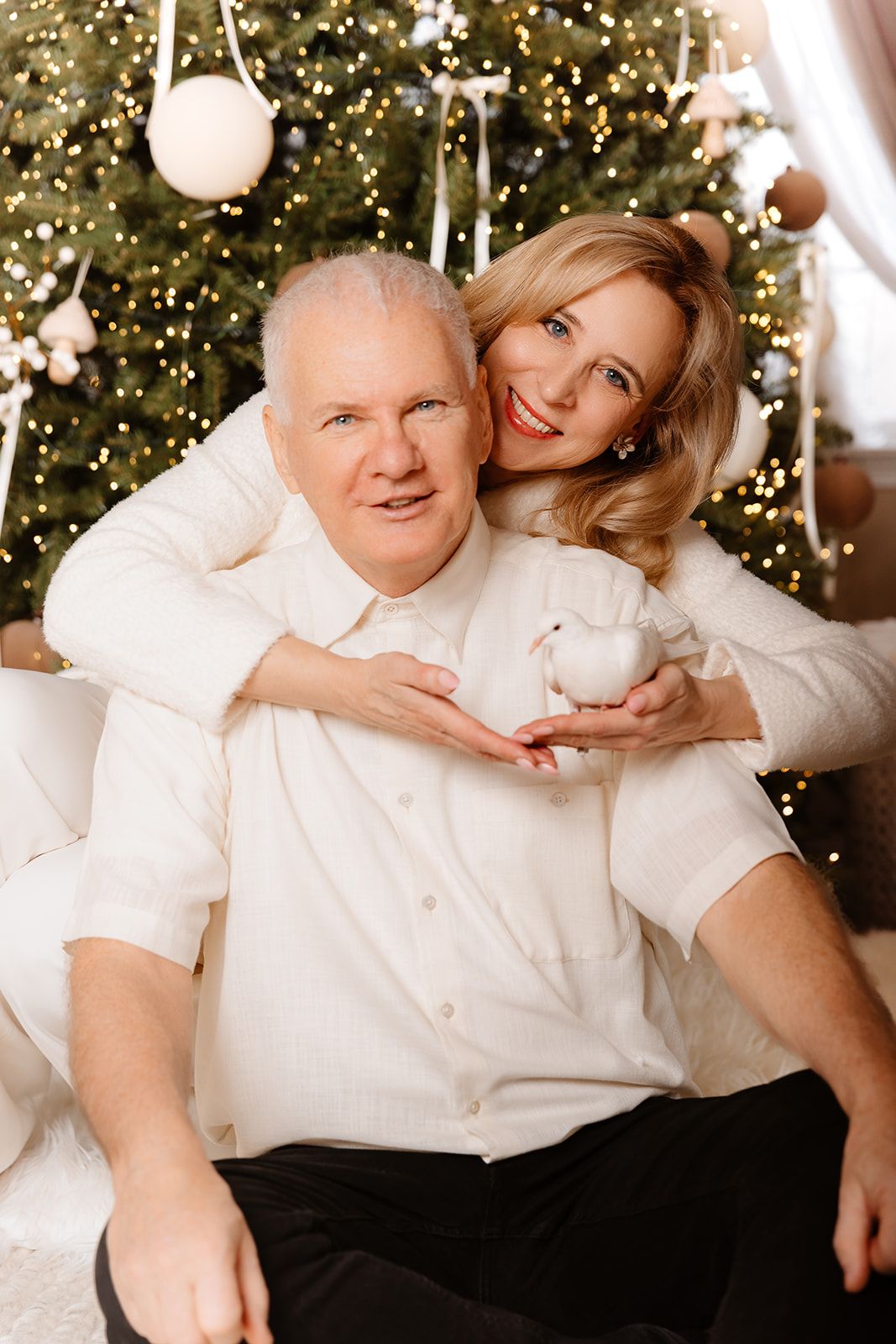 Happy couple in white sweaters embracing near decorated Christmas tree, woman holding white dove ornament.