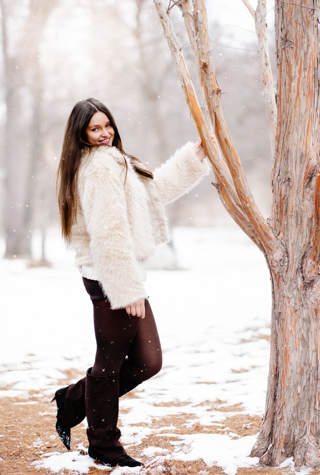 A teenage girl in a white faux fur coat and dark boots leans against a tree trunk in a snowy winter forest.