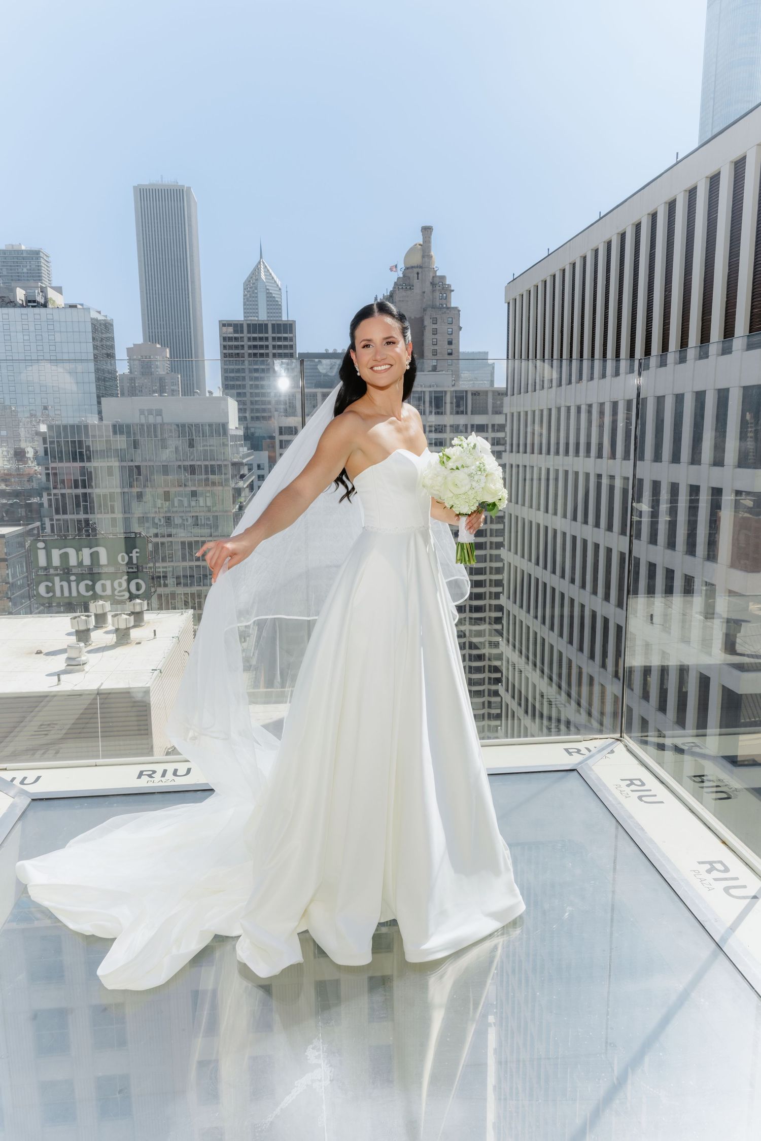 Radiant bride in white gown and flowing veil posing on glass rooftop with Chicago skyline.