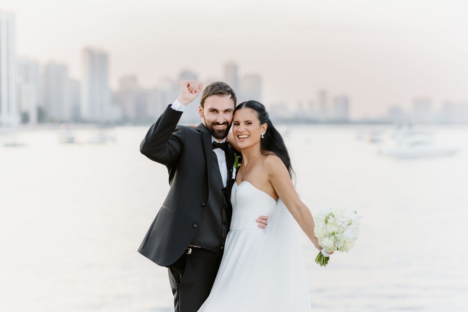 Smiling bride and groom celebrate by the water with a city skyline behind them on their wedding day.