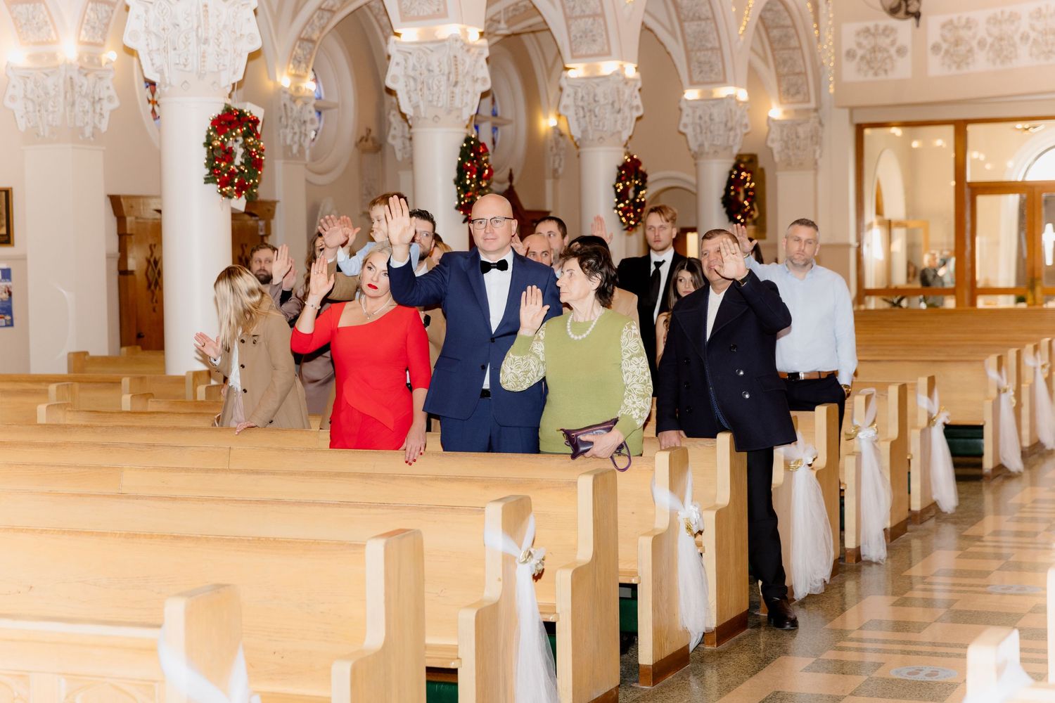 Wedding guests wave and smile as they walk down the aisle of an ornate church decorated for Christmas.