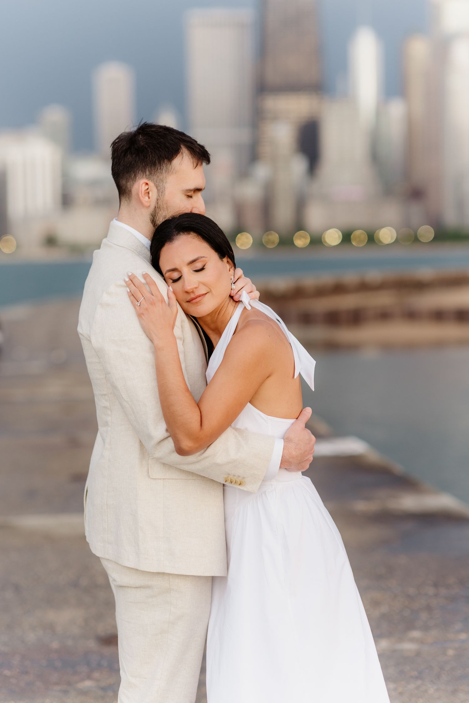 Couple shares tender embrace on pier as man kisses womans head with Chicago skyline bokeh backdrop.