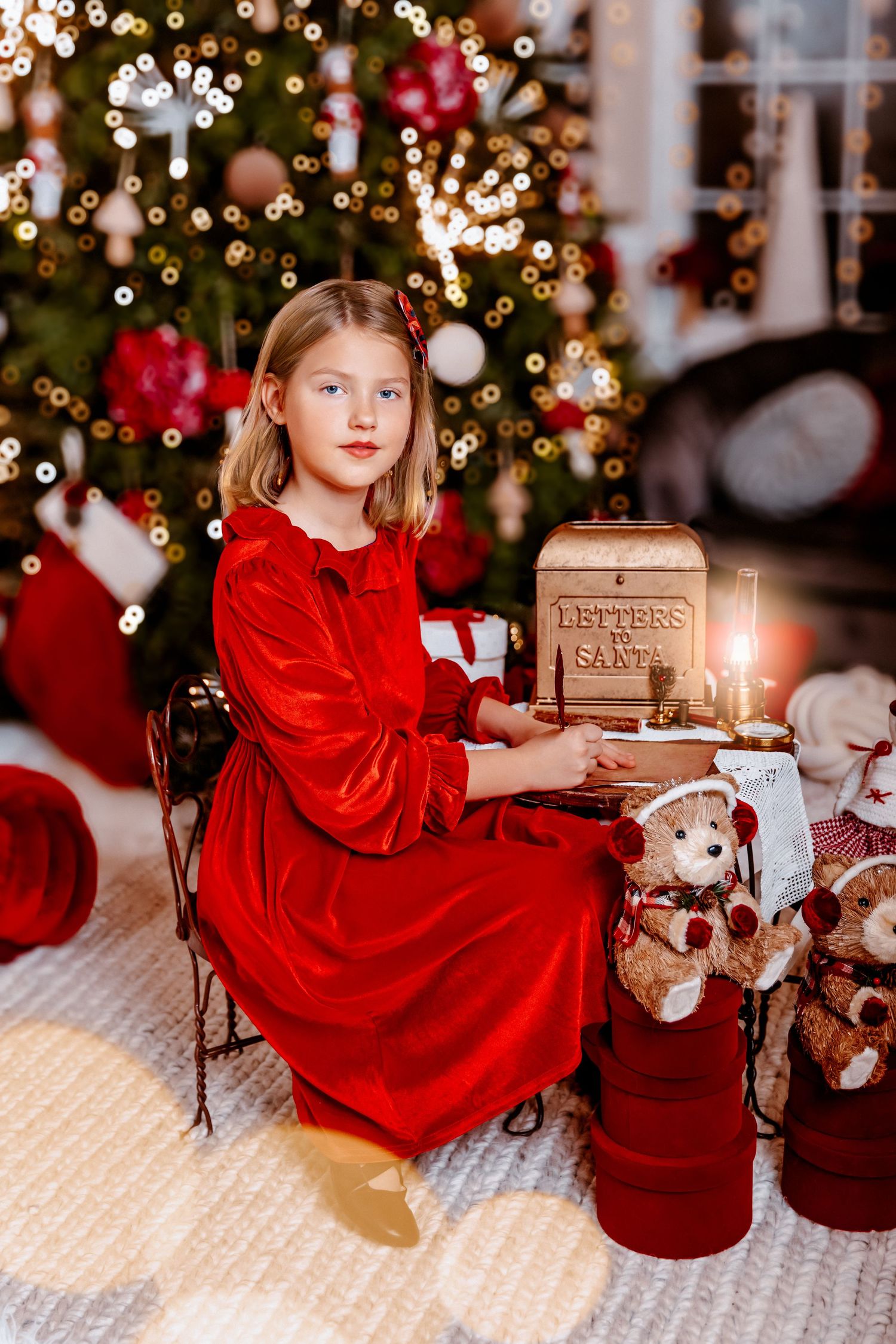 Young girl in red velvet dress writing letter to Santa beside Christmas tree with teddy bears and candle.