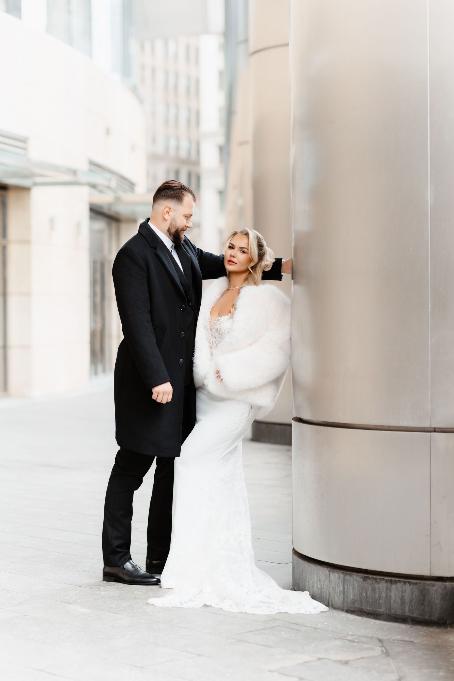 Bride in white lace gown with fur wrap leans against column as groom gazes at her downtown.