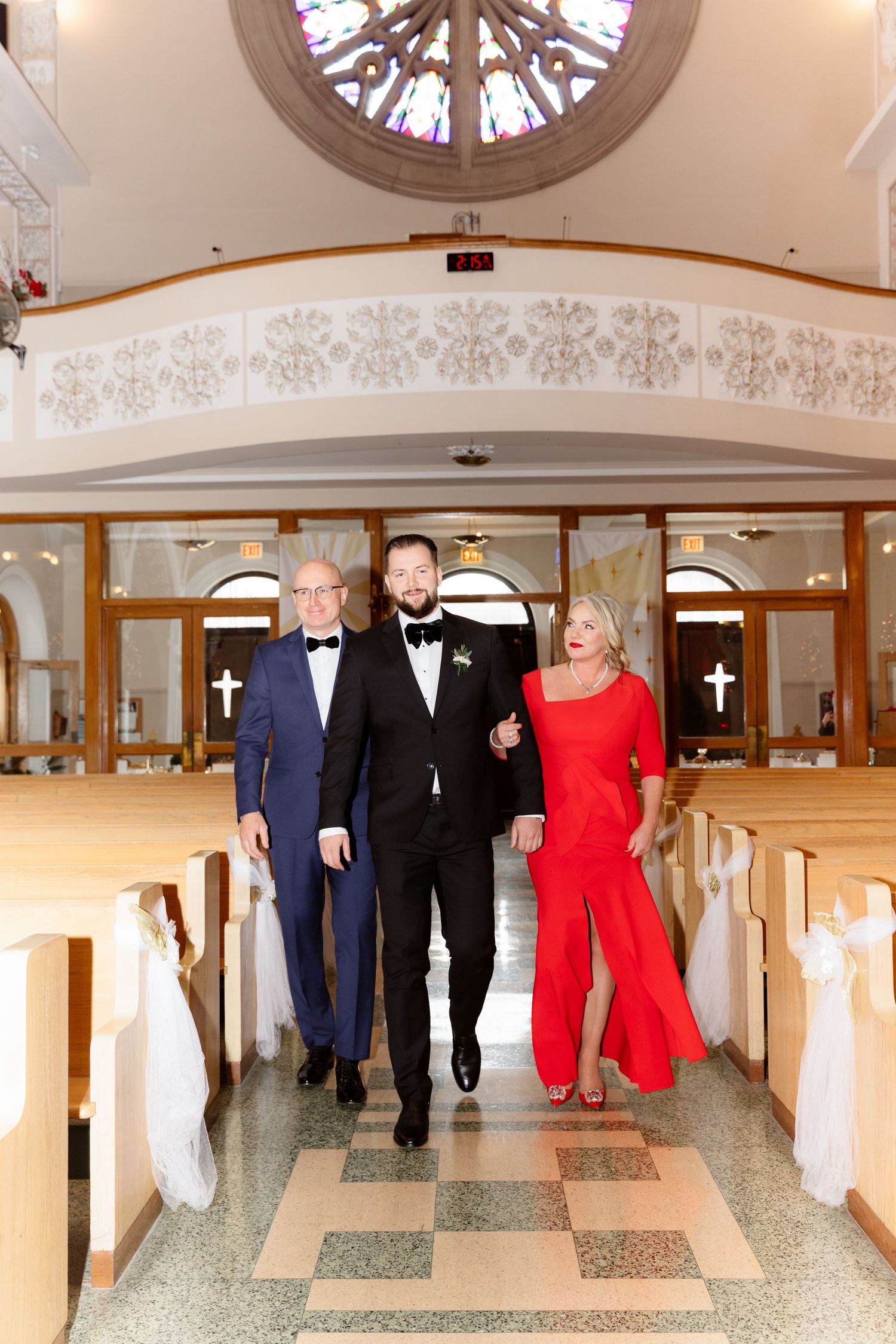 Groom in black tuxedo walks down church aisle with parents during wedding ceremony processional.