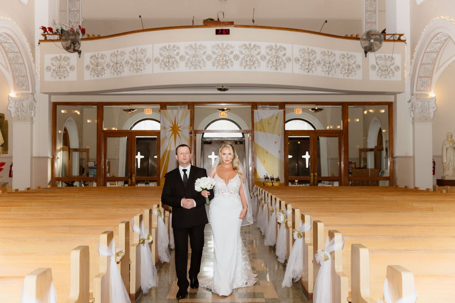 Bride and groom walking down the aisle of an elegant white church with ornate decor during wedding ceremony.