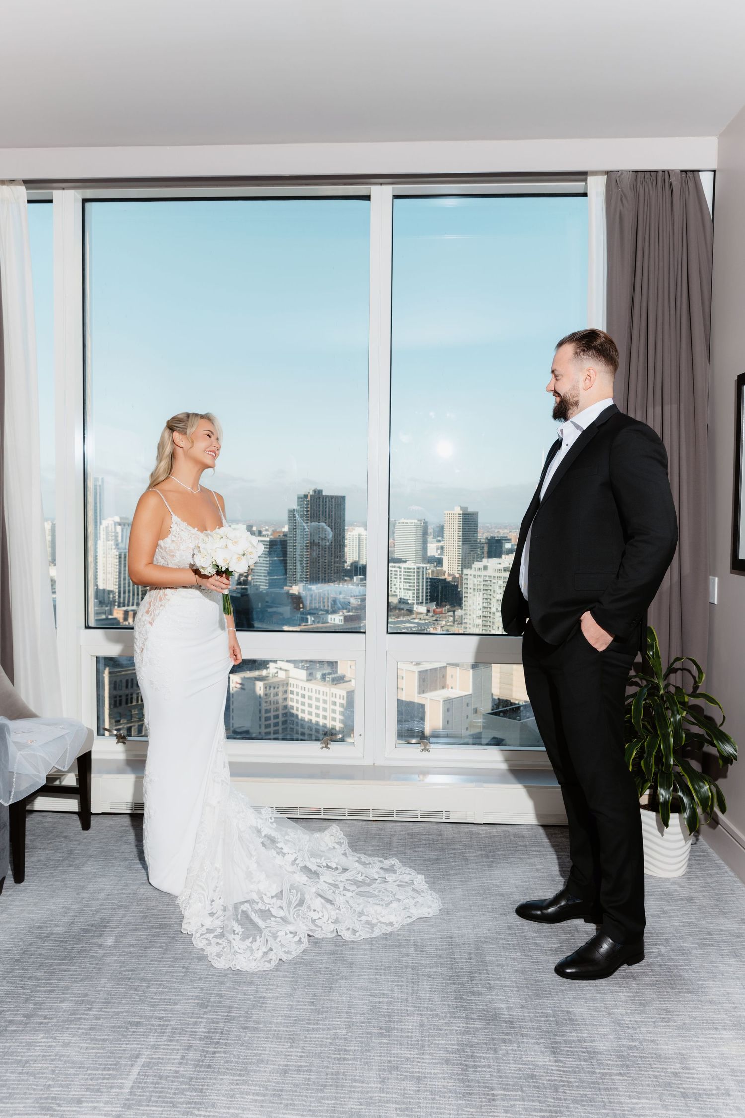 Bride and groom share a first look moment in a sunlit hotel suite with city skyline backdrop.