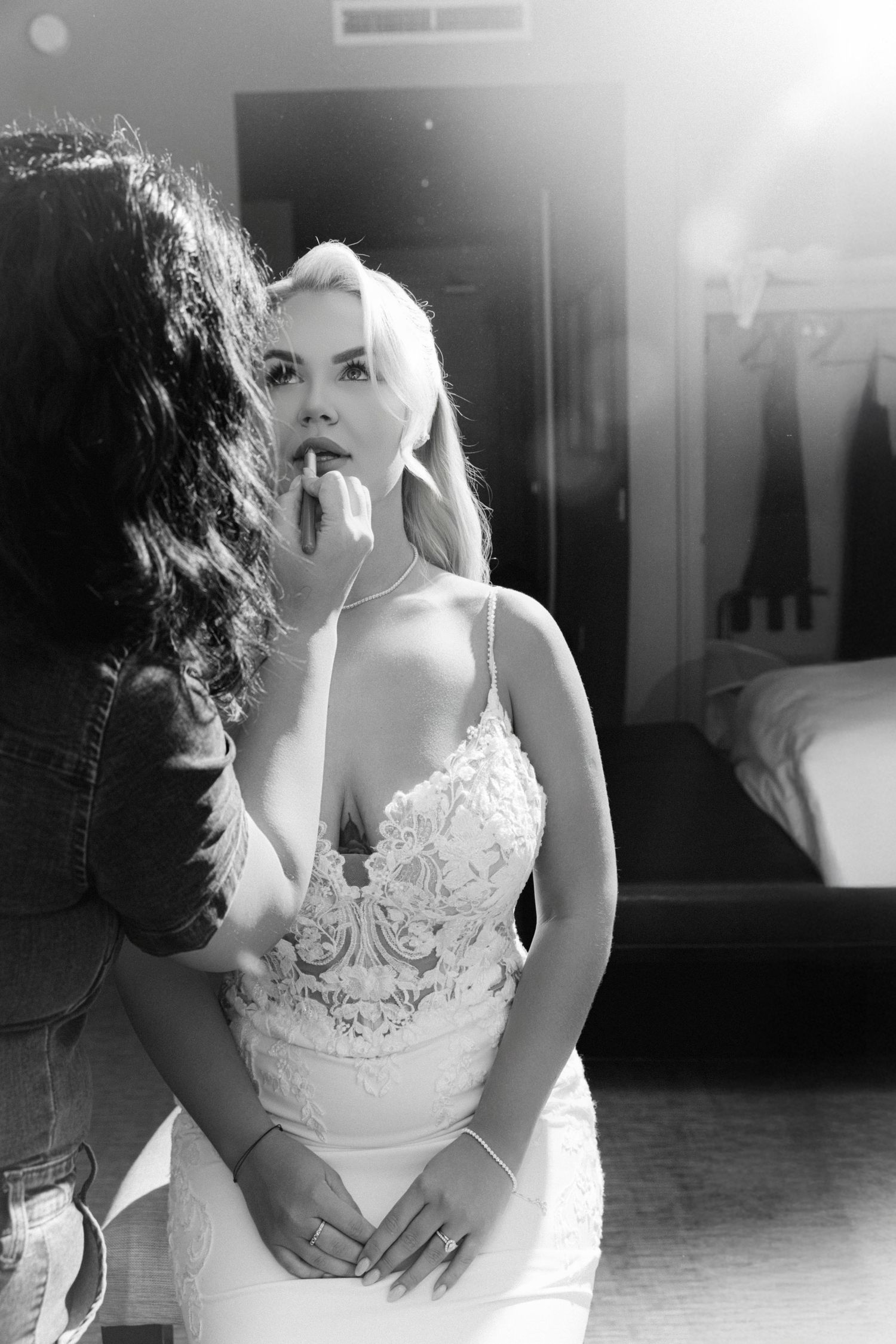 Black and white photo of makeup artist applying lipstick to blonde bride in lace wedding dress.