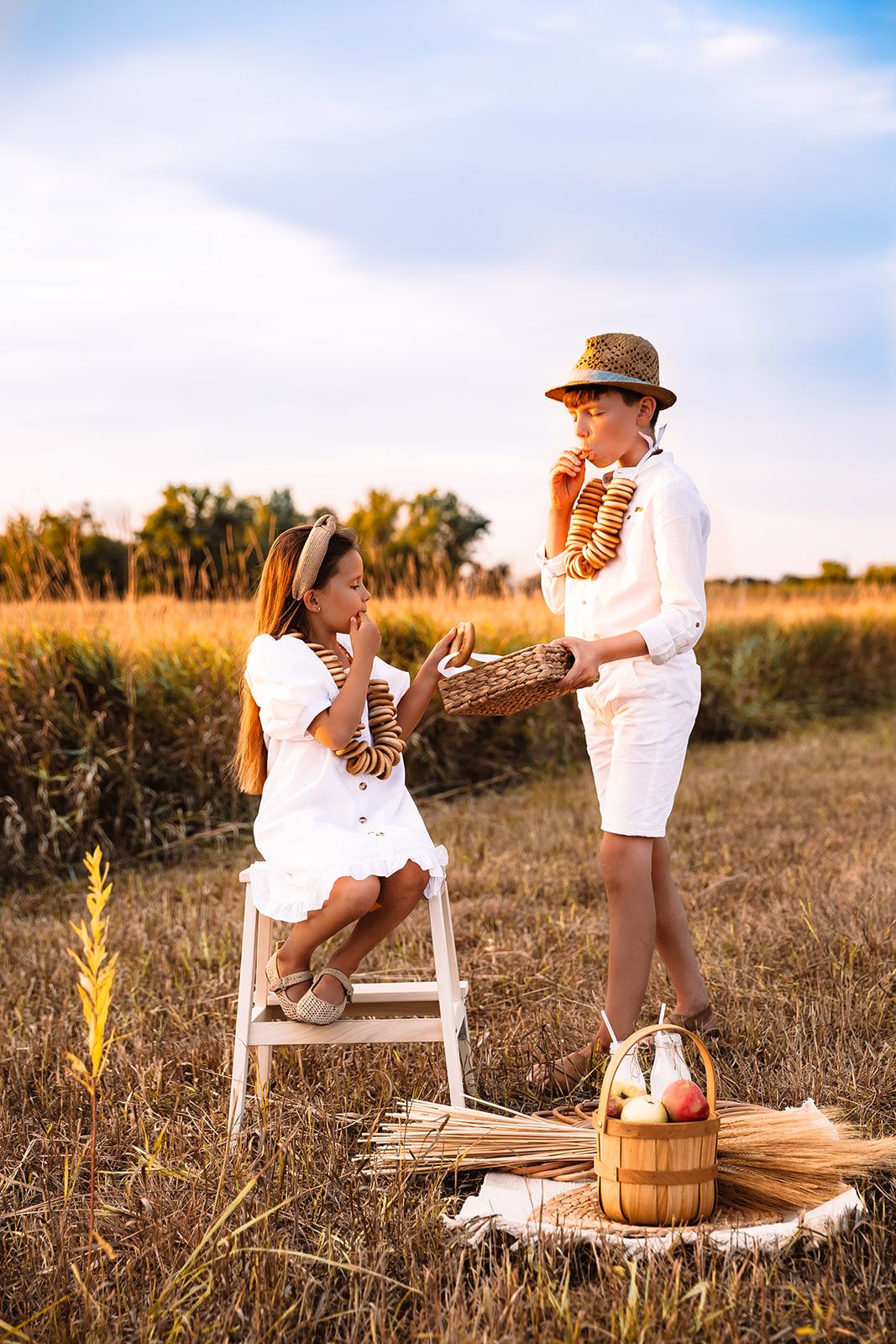Two children in white outfits share bread in a sunny field, with a basket and apples nearby.