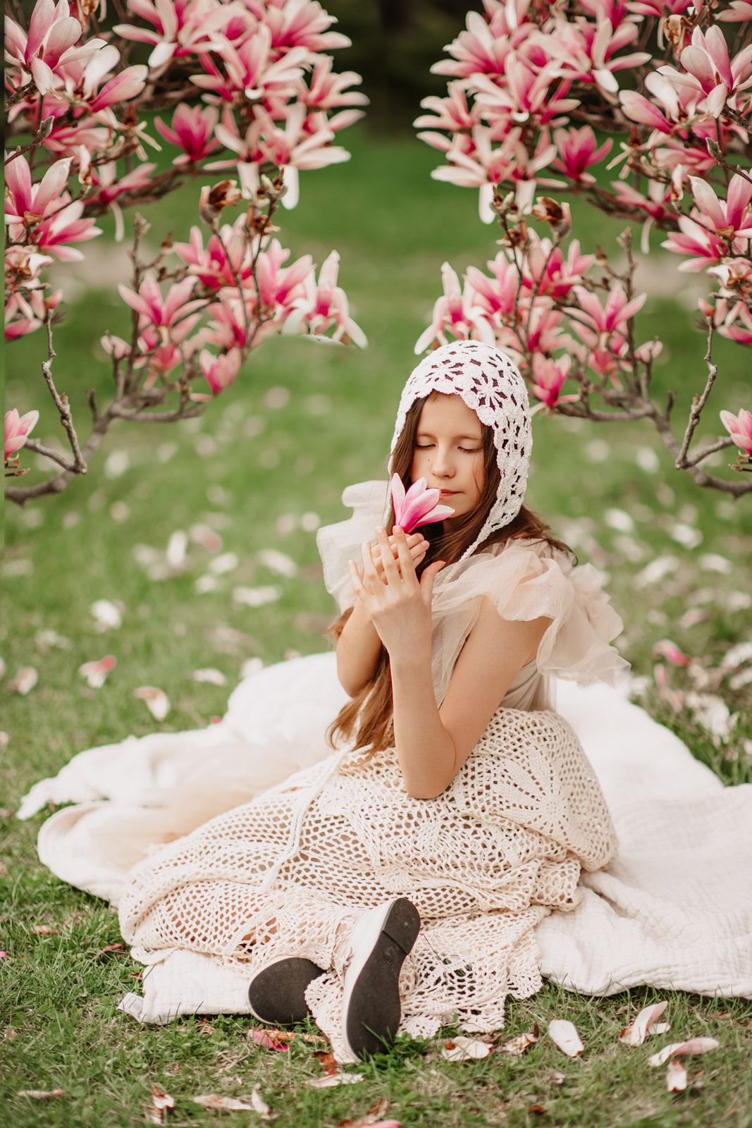 Young girl in cream crochet dress and bonnet sits on blanket smelling pink magnolia flower under blooming tree.