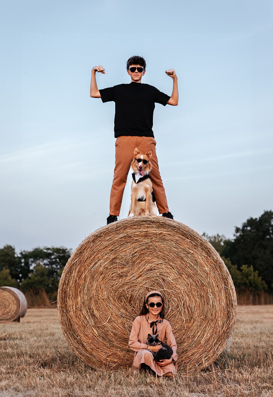 Boy flexing on hay bale with dog while girl sits below in sunny farm field.