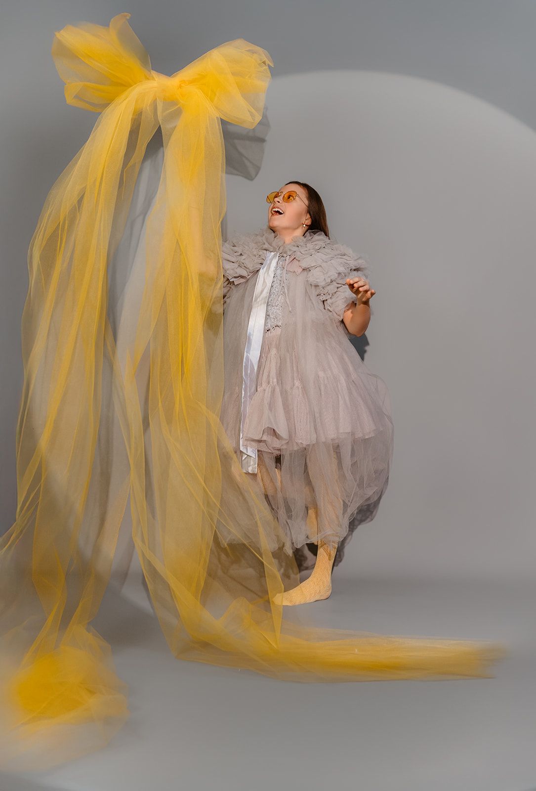 Woman in gray ruffled dress with yellow tulle bow and veil, wearing yellow sunglasses, posing on gray background.
