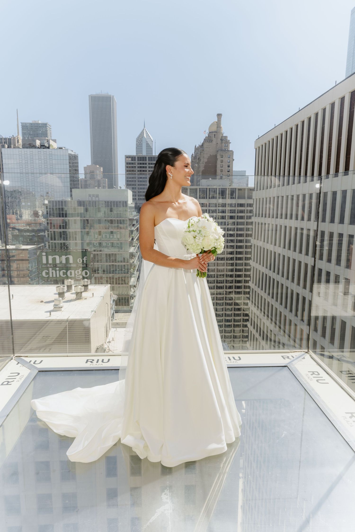 Bride in strapless white gown holding white bouquet standing on rooftop with Chicago skyline.