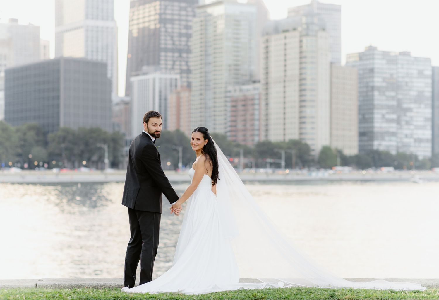 Elegant bride and groom posing by lakefront with stunning city skyline behind them.