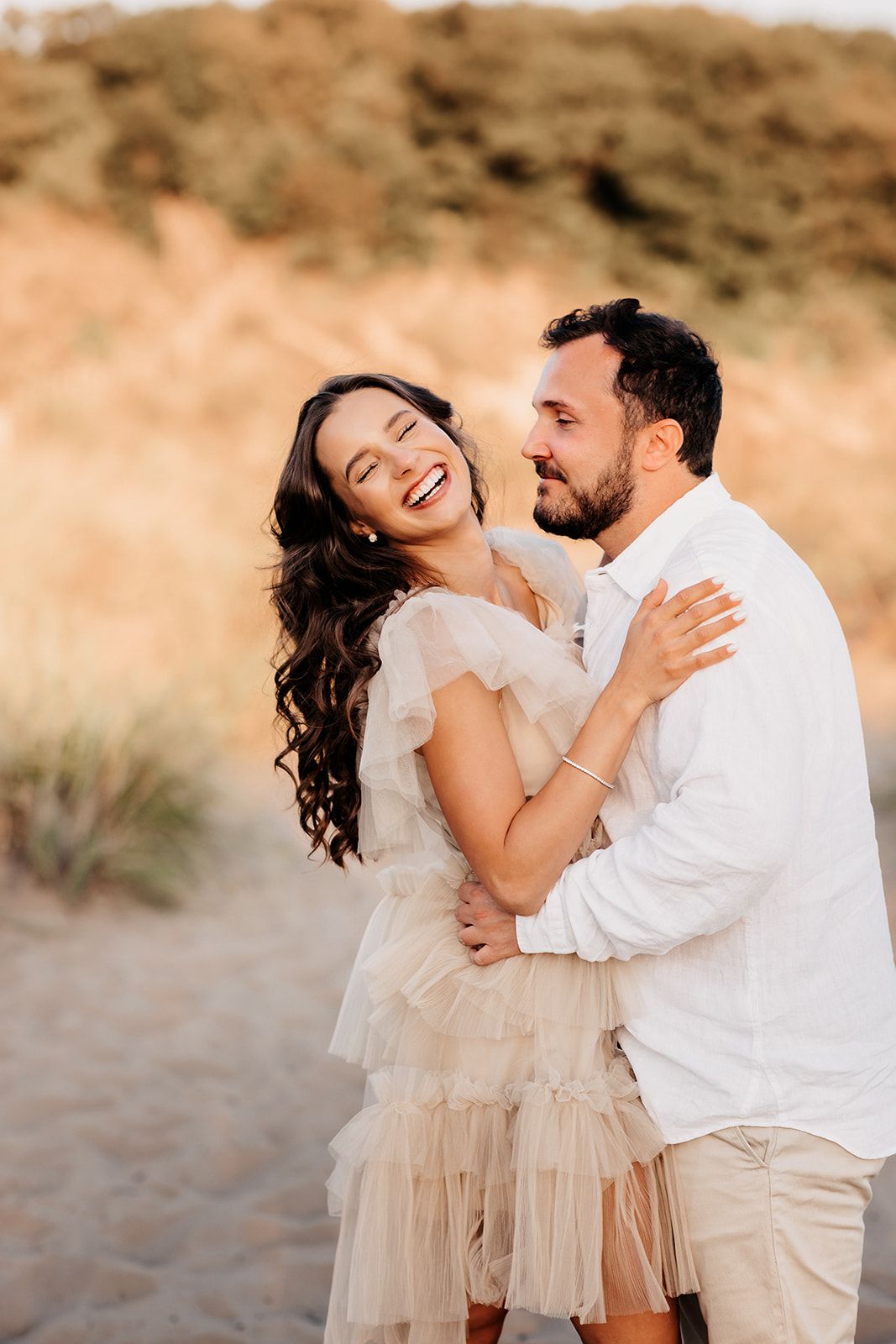 Happy couple embracing and laughing on sandy beach path with golden grass background.
