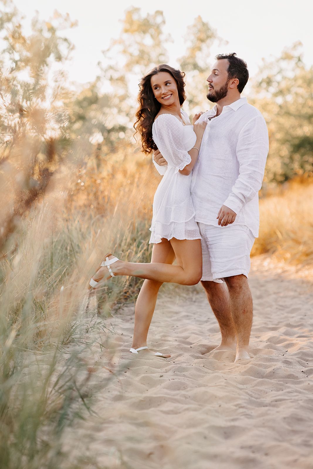Happy couple in matching white outfits embrace on sandy beach path at golden sunset.