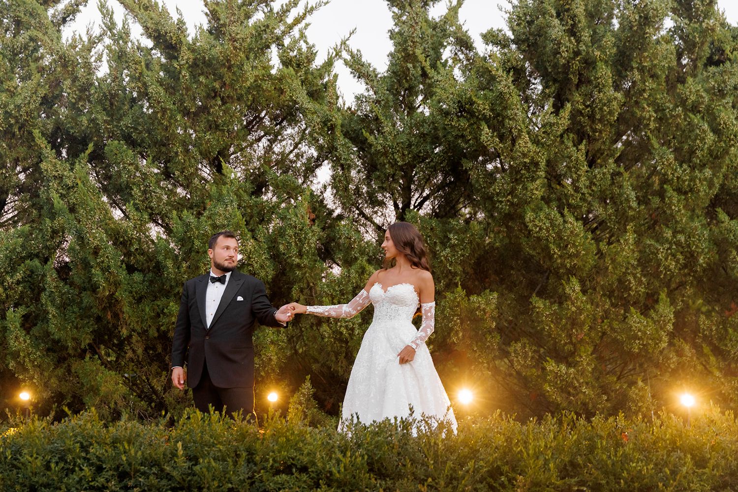 Bride and groom hold hands in garden at dusk, surrounded by evergreen trees and soft glowing lights.