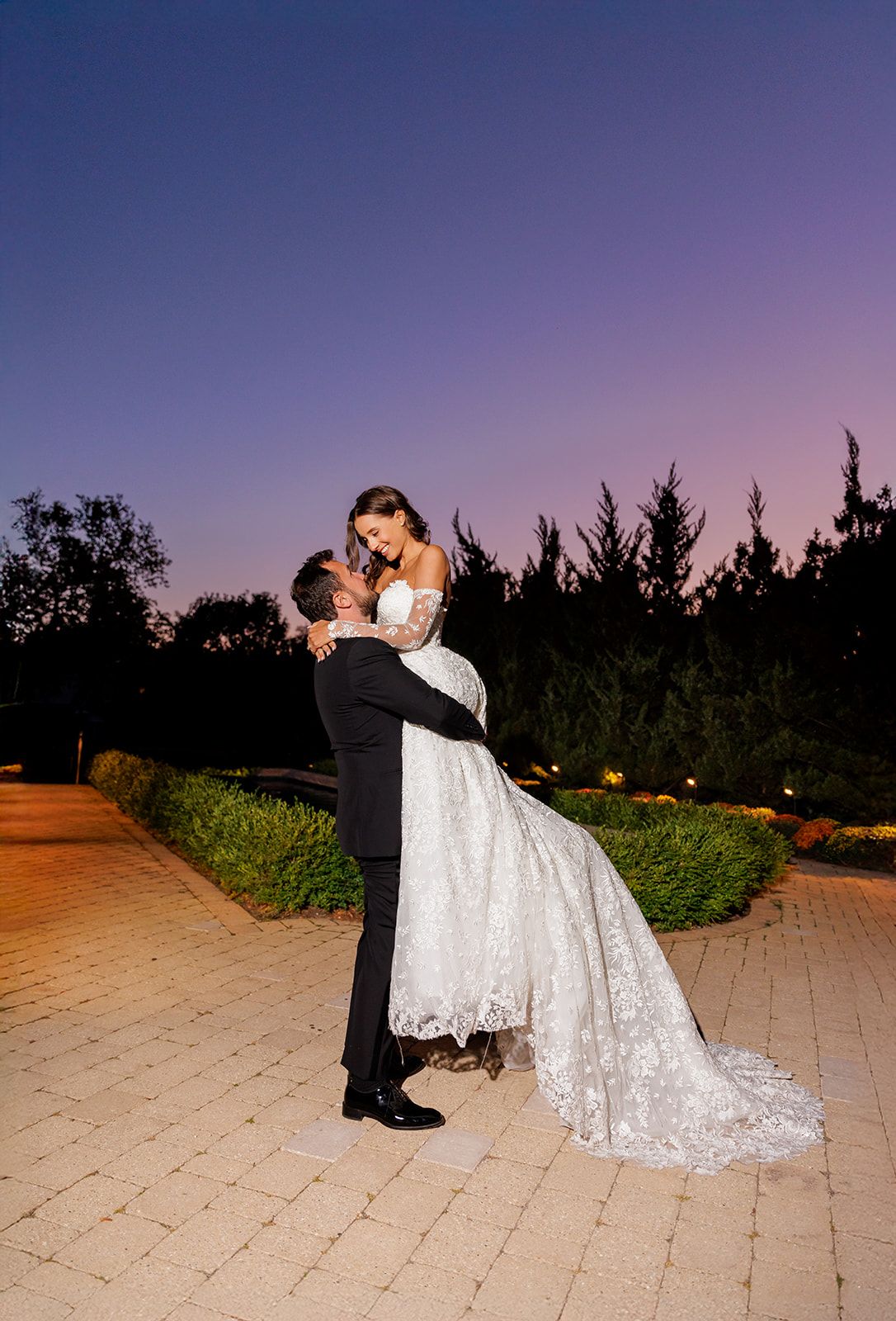 Groom lifts bride in lace wedding gown at twilight, sharing a romantic kiss against a purple sunset sky.