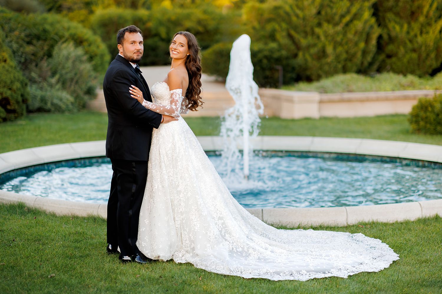 Bride and groom stand elegantly by circular garden fountain, bride wearing lace gown with cathedral train.