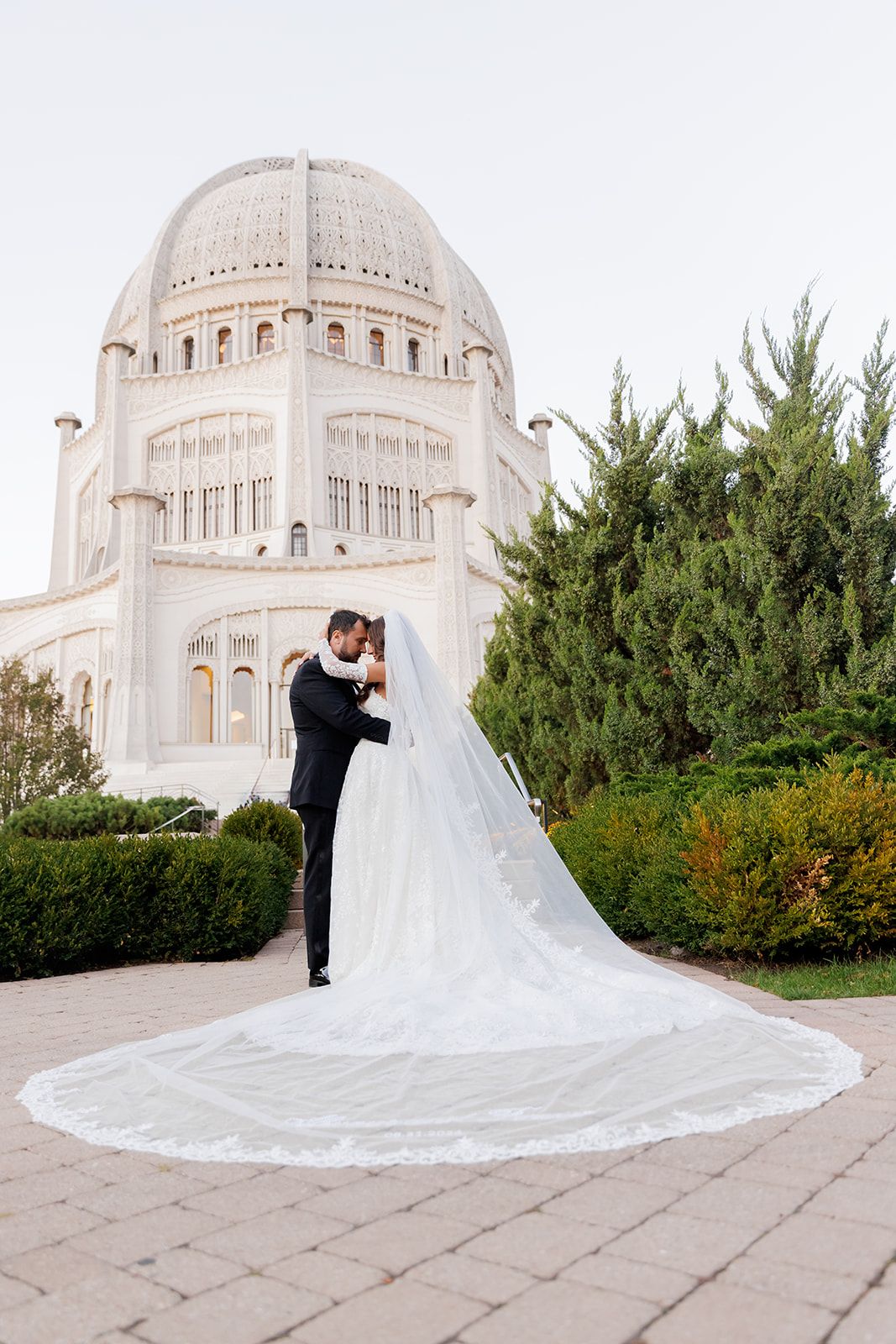 Bride and groom embracing before a white domed Bahai Temple, bride's long lace veil spreading elegantly on stone pavement.
