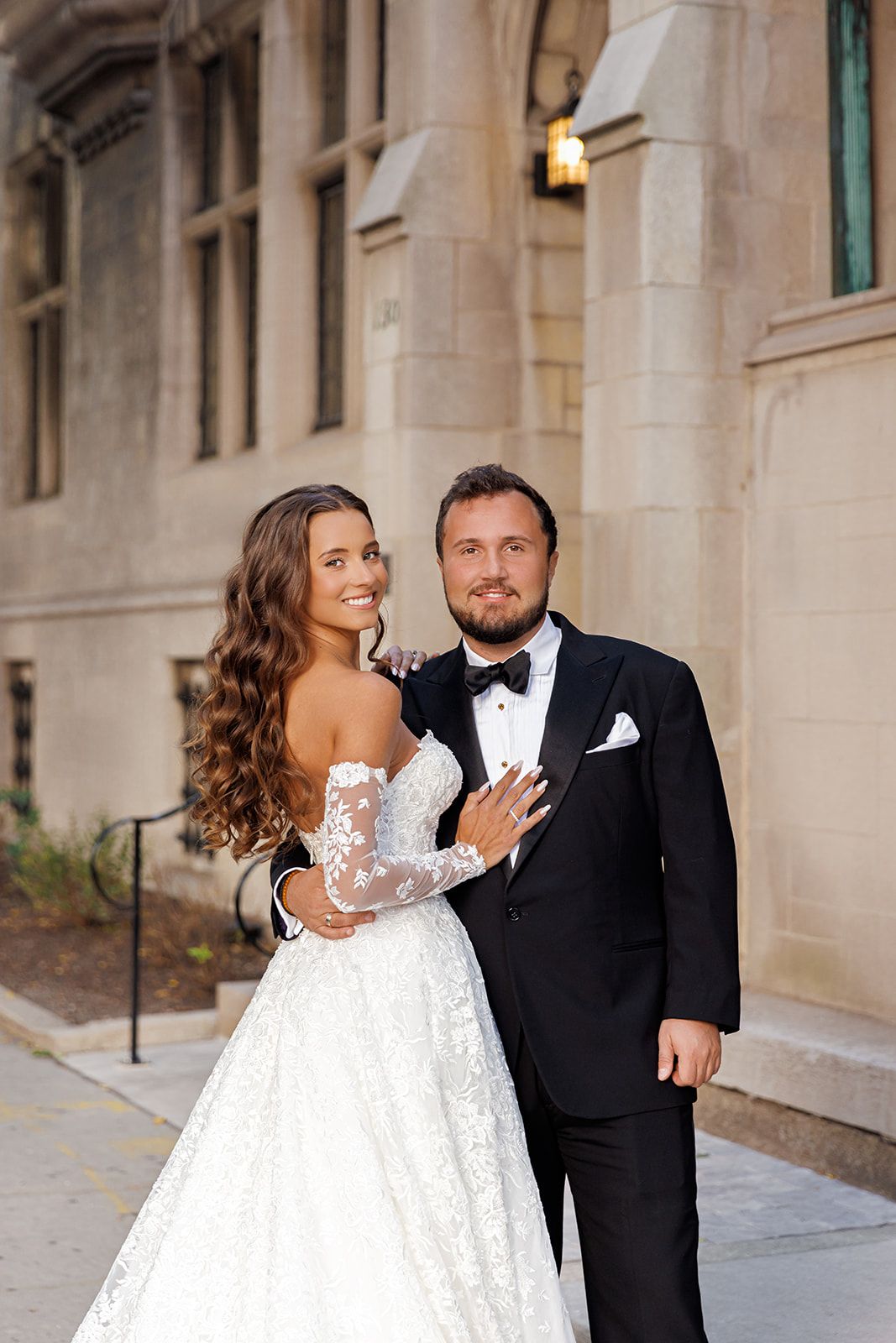 Bride in lace off-shoulder gown embraced by groom in black tuxedo, posing outdoors near elegant stone building.