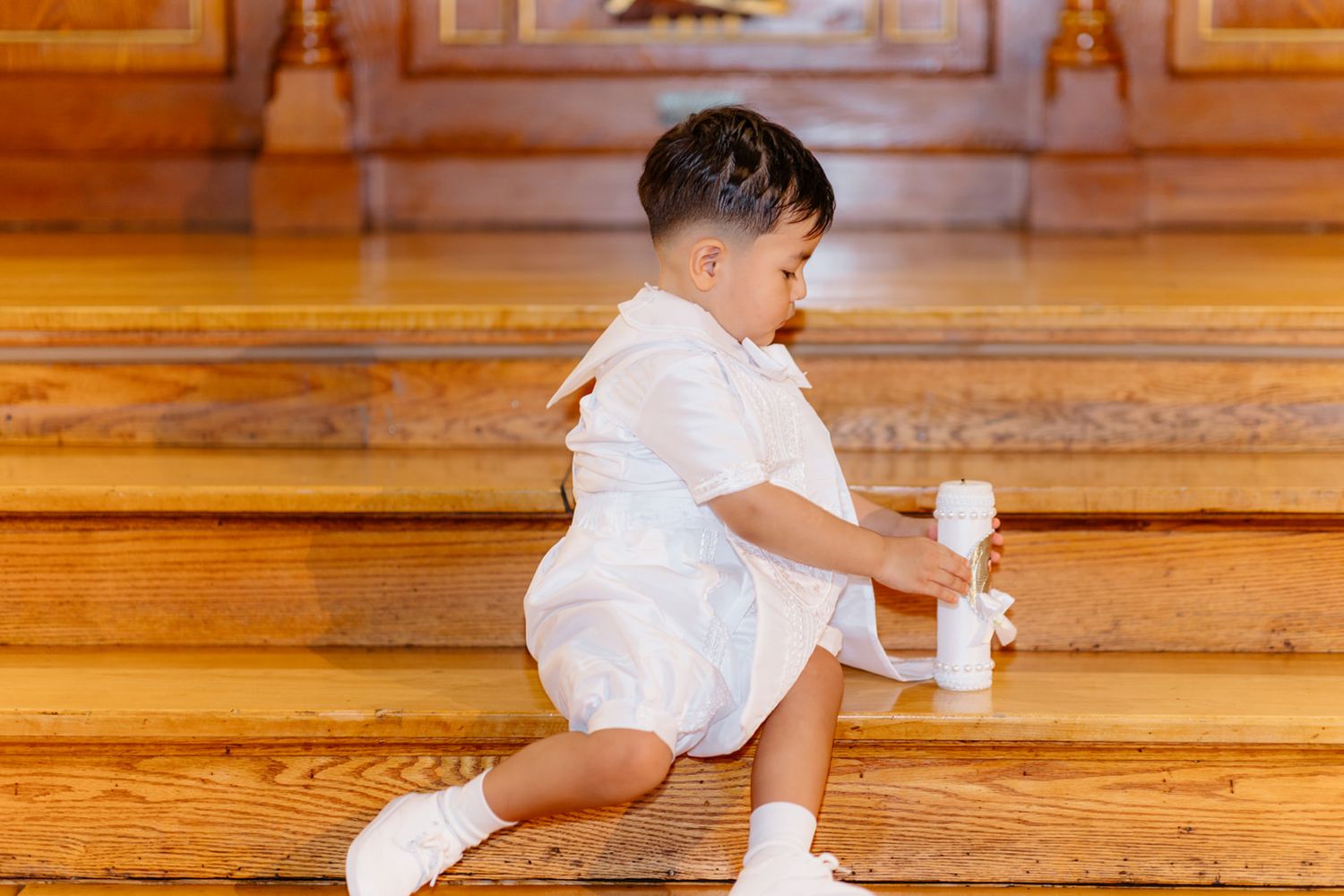 Toddler boy in white outfit sitting on wooden church steps holding a baptism candle.
