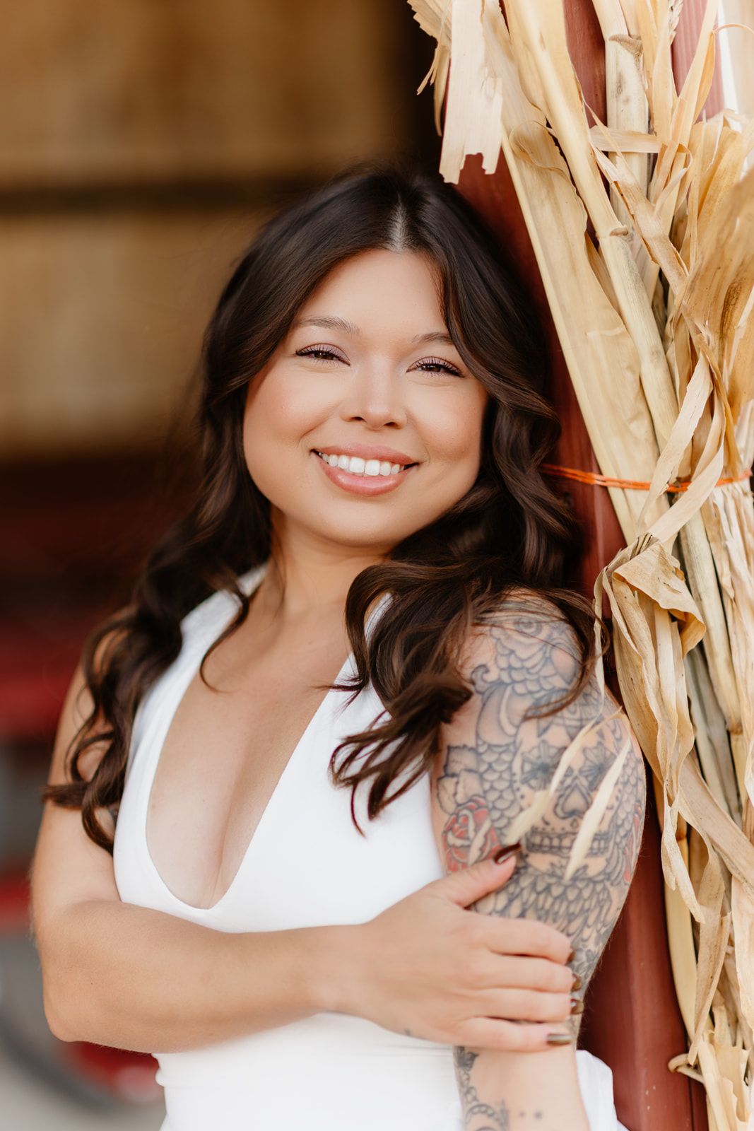 A smiling bride in a white dress with a floral tattoo sleeve leans against a barn decoration.