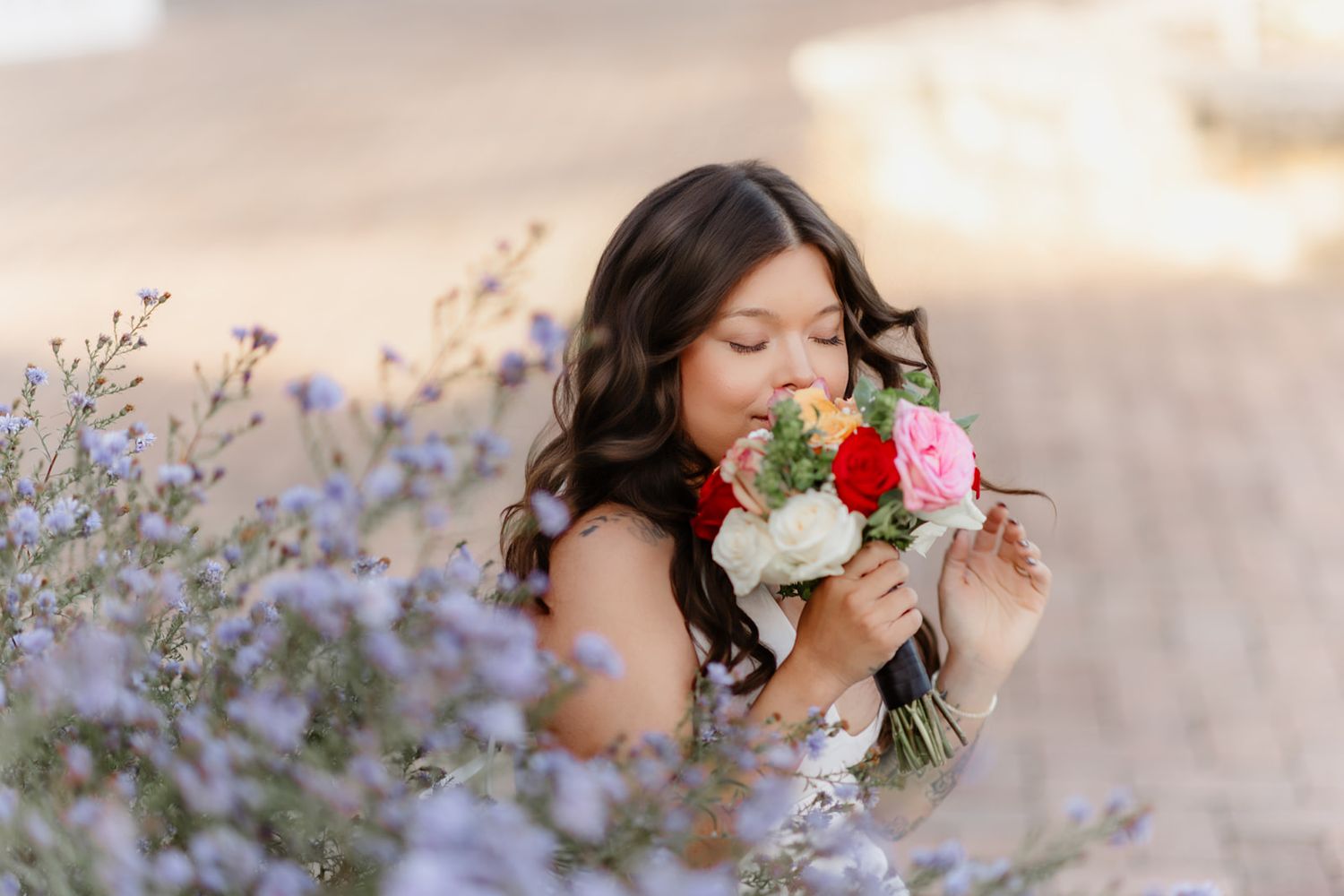Young woman in white dress smells colorful bridal bouquet surrounded by purple wildflowers in soft golden light.
