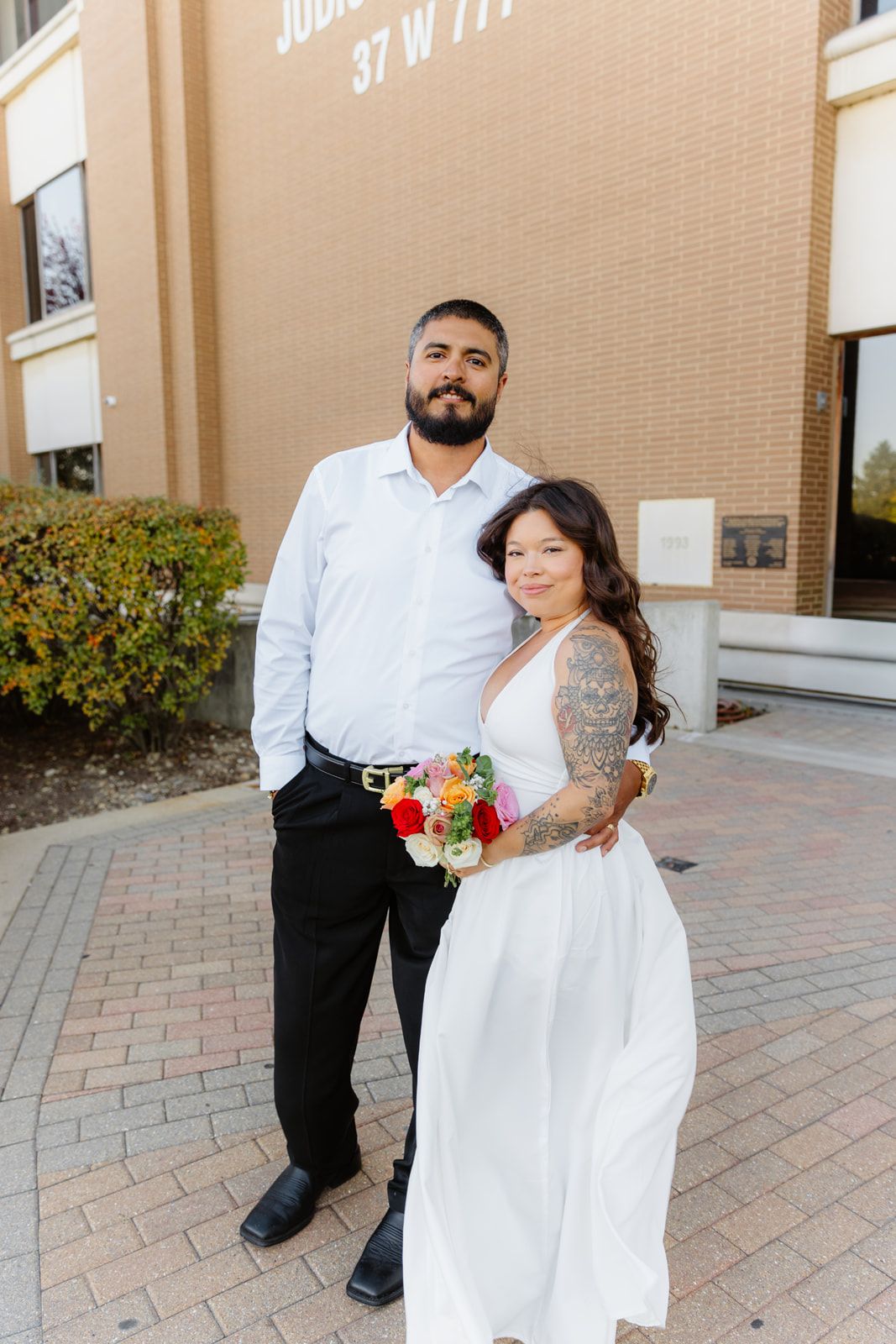 Couple poses in wedding attire outside Kane County Judicial Center, bride holding colorful bouquet.