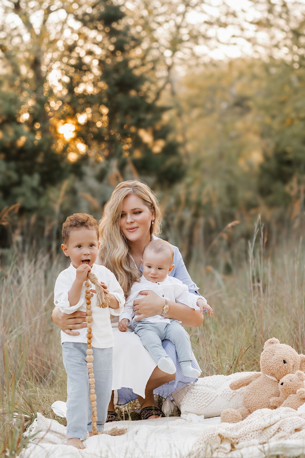 A blonde mother sits in a golden field holding her baby while her toddler stands beside her with a teddy bear.