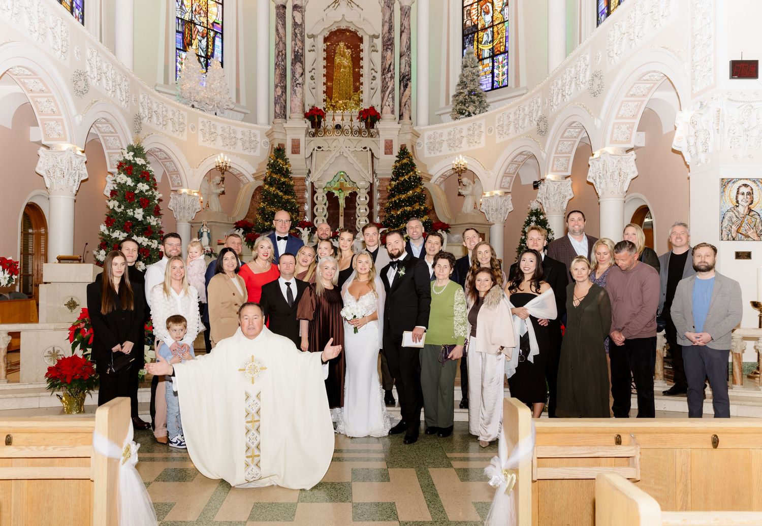 Large wedding group photo with priest in white vestments inside beautifully decorated Christmas church ceremony.