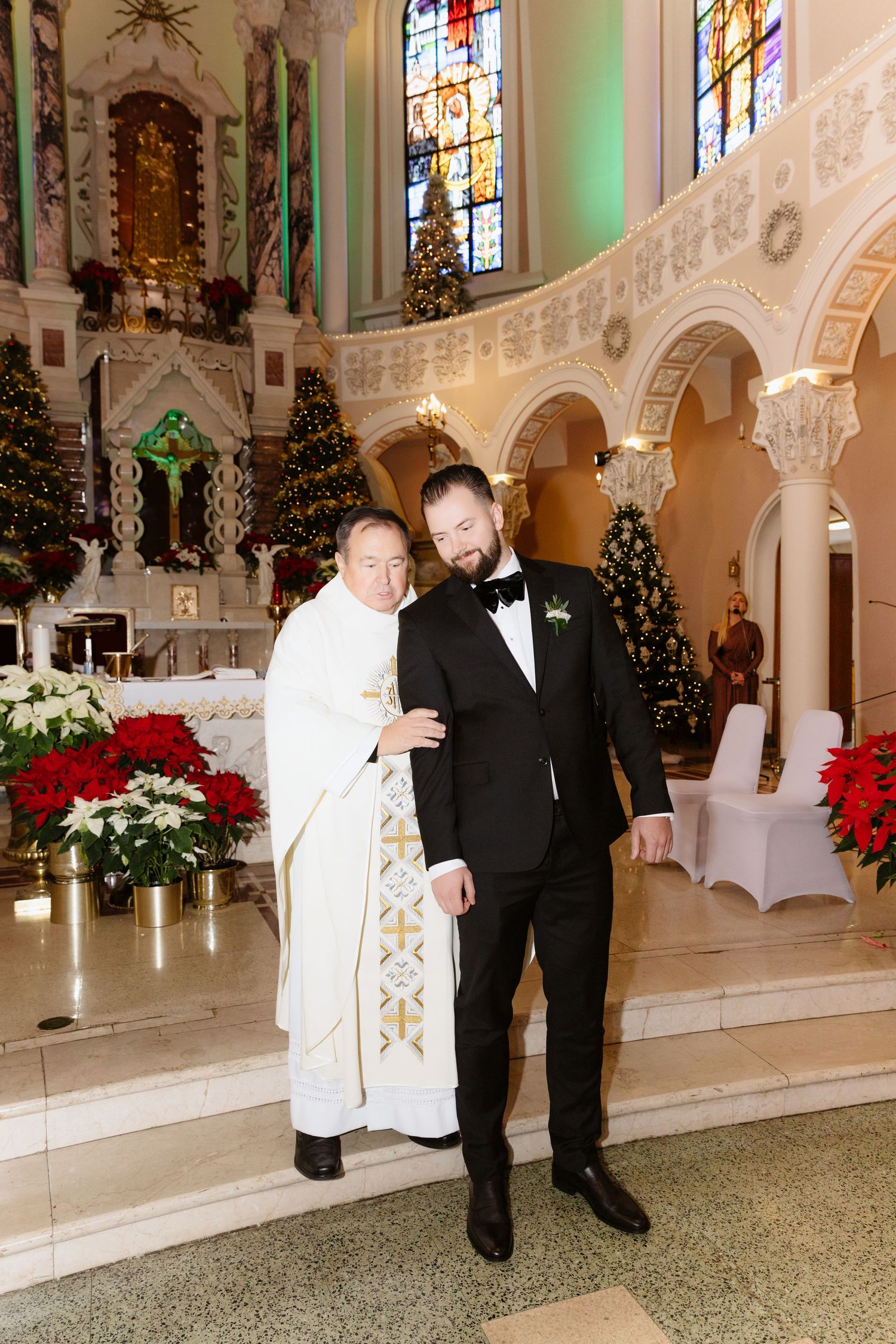 Groom in tuxedo poses with priest in white vestments at decorated church altar with Christmas trees.