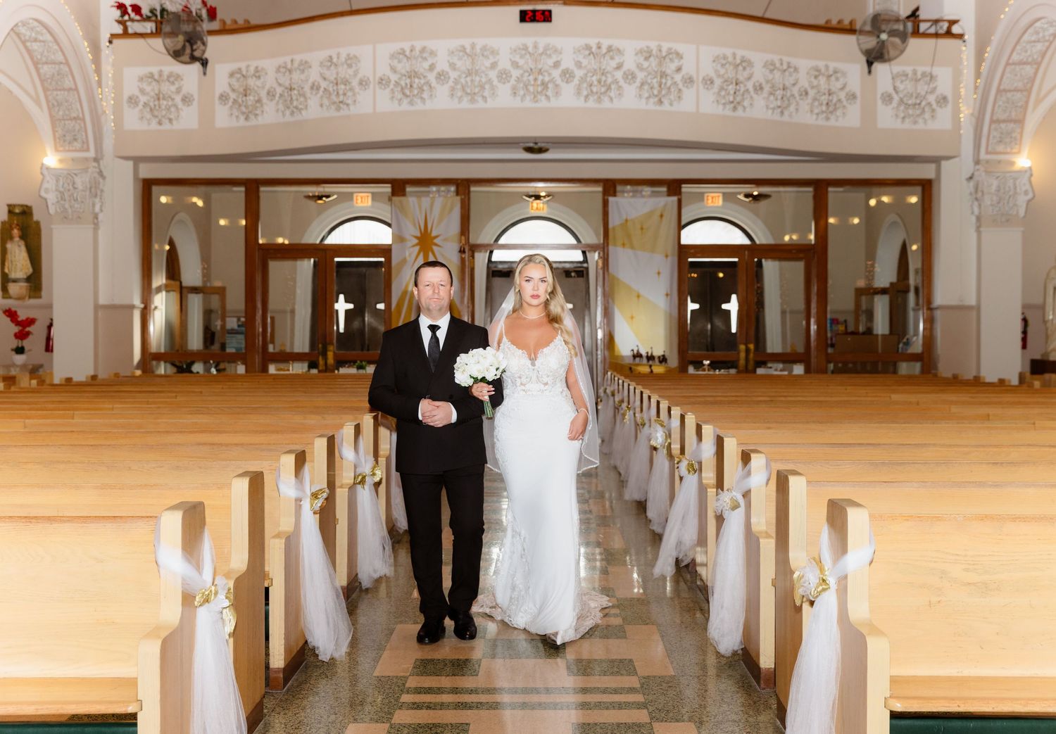 A bride and escort walk down a church aisle lined with wooden pews and white floral decorations.