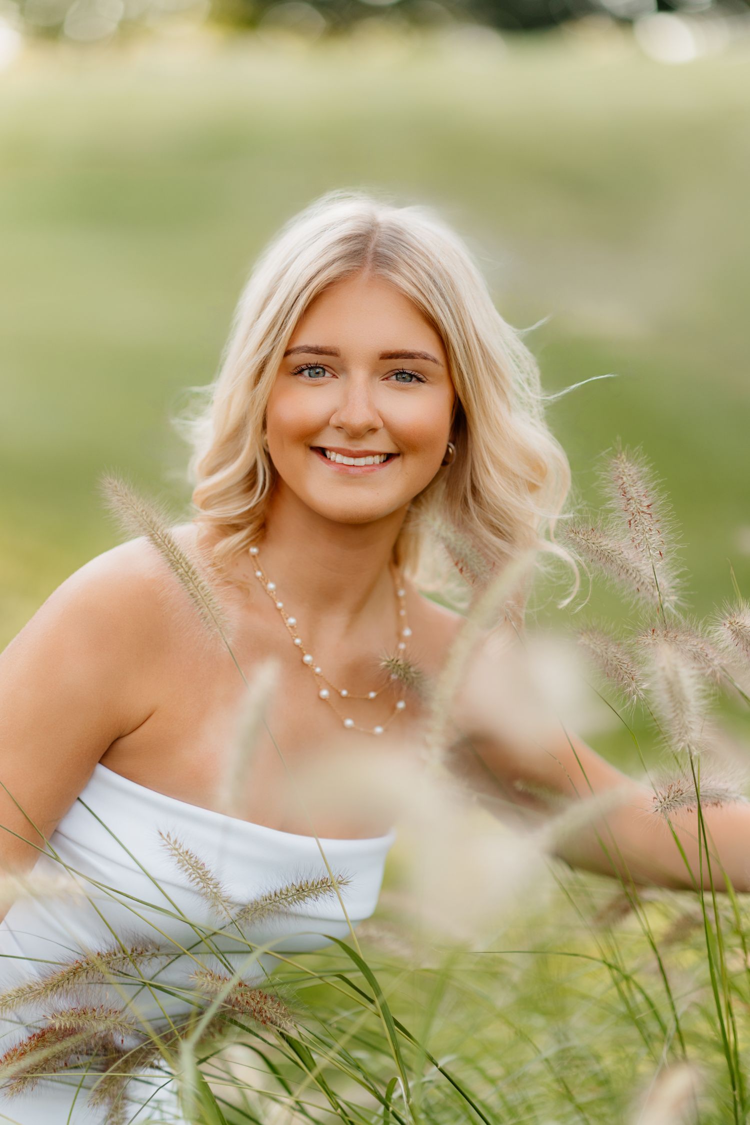 Smiling blonde woman in white dress surrounded by tall ornamental grasses outdoors.