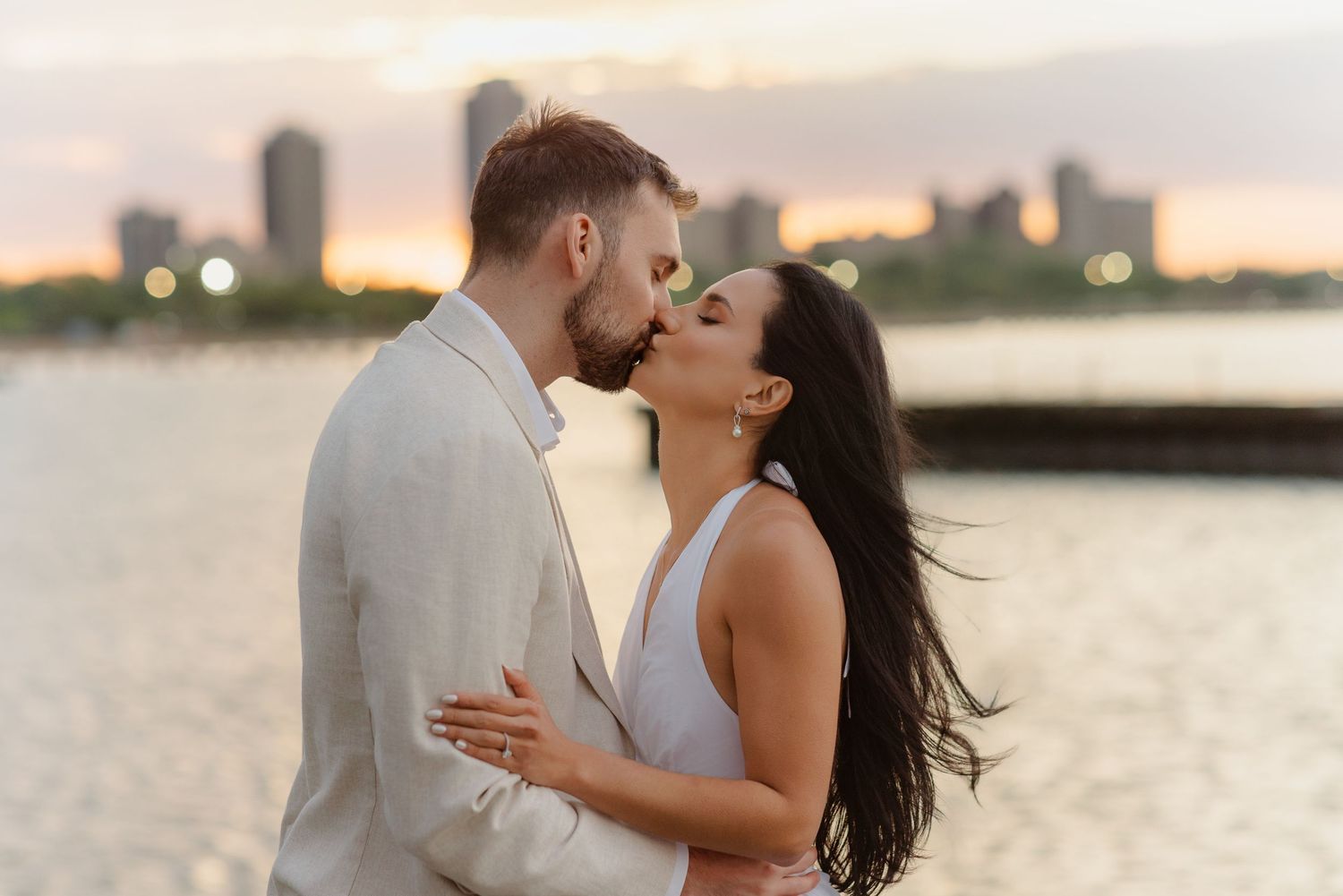 Couple sharing romantic kiss by waterfront with colorful sunset city skyline behind them.
