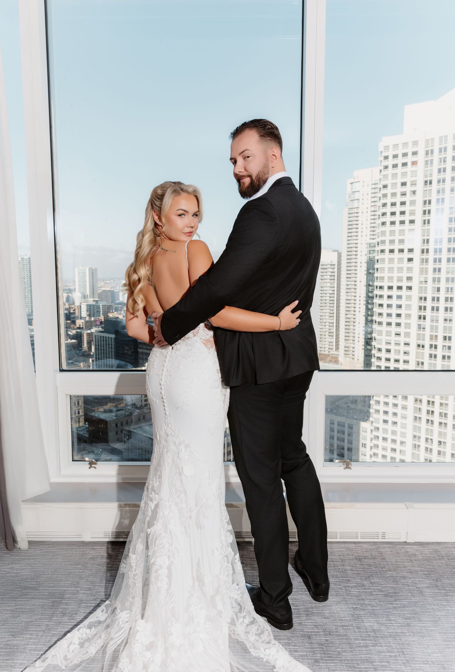 Bride in backless lace mermaid gown posing with bearded groom in black tuxedo against city skyline.
