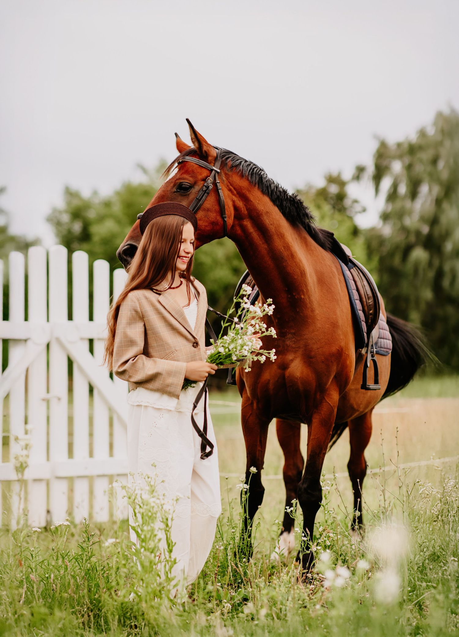 Woman in beige blazer offers wildflowers to a tall chestnut horse standing by a white picket fence.